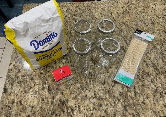 photo of a bag of sugar, some jars, sticks, and food coloring on a counter